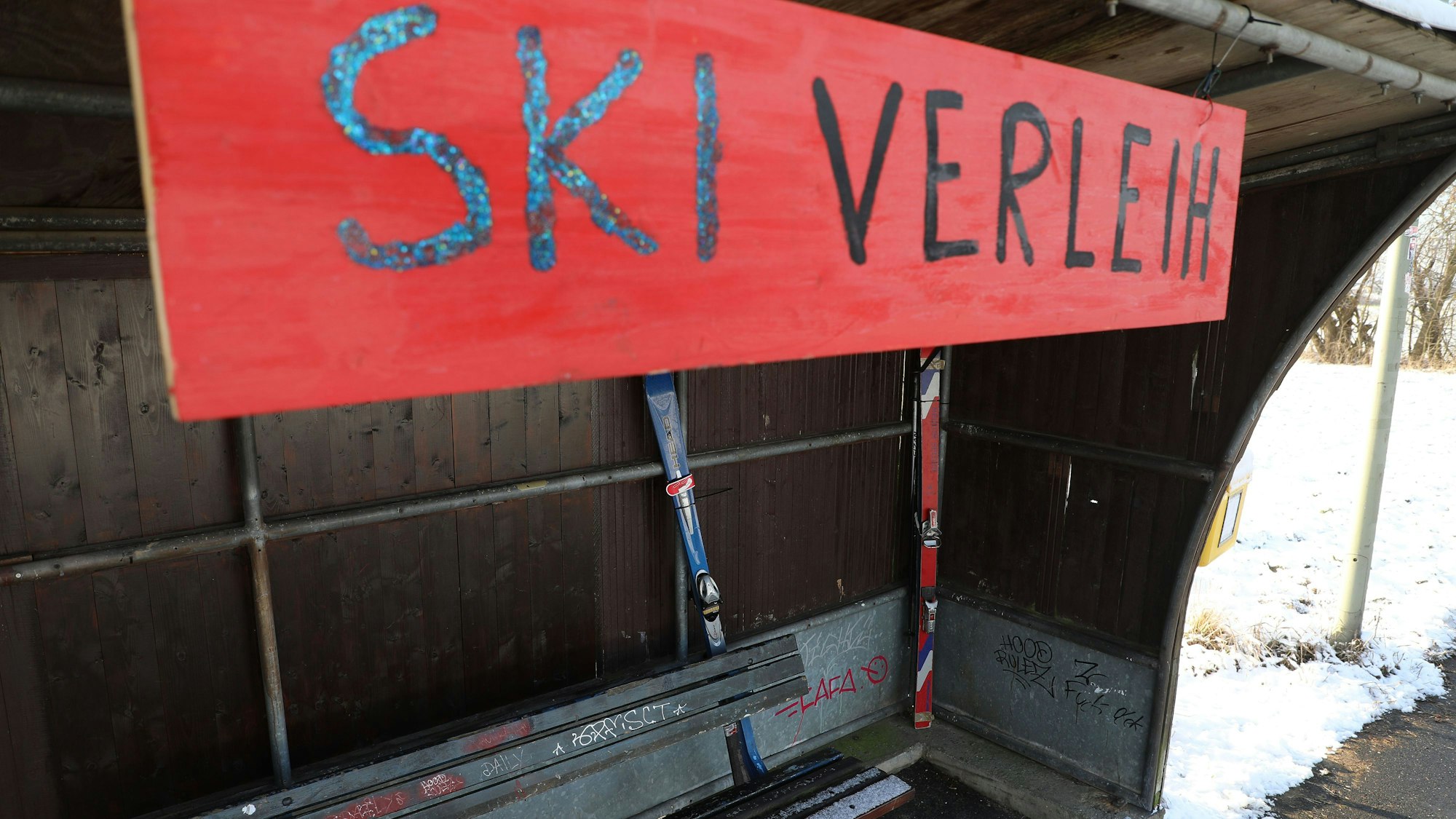 An einer Bushaltestelle hängt ein Schild mit der Aufschrift „Ski Verleih“ im Inneren des Wartehäuschens sind Ski zu sehen.