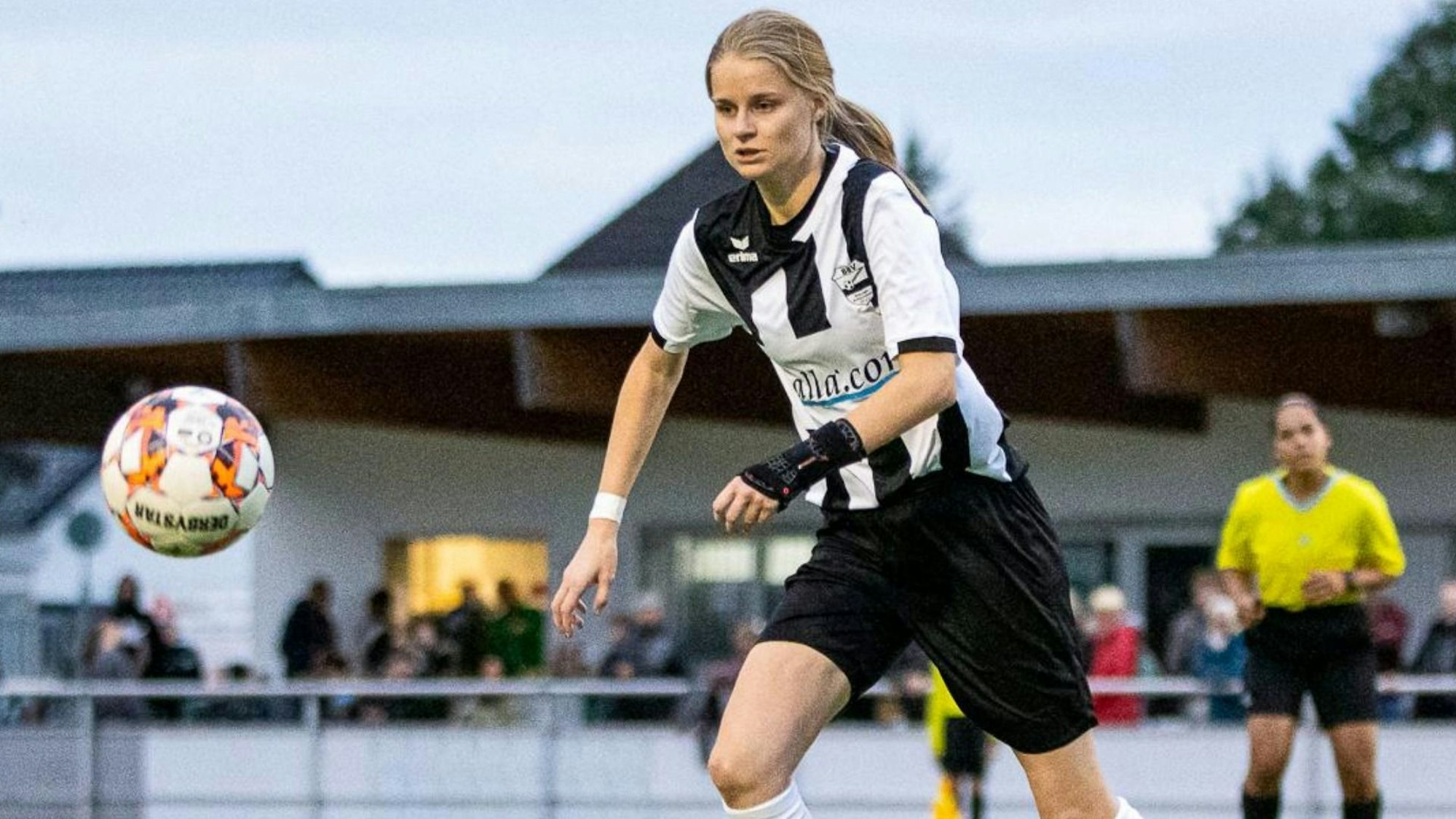 Eine Fußballspielerin läuft über einen Platz, vor ihr ist ein Fußball zu sehen, im Hintergrund eine Schiedsrichterin.