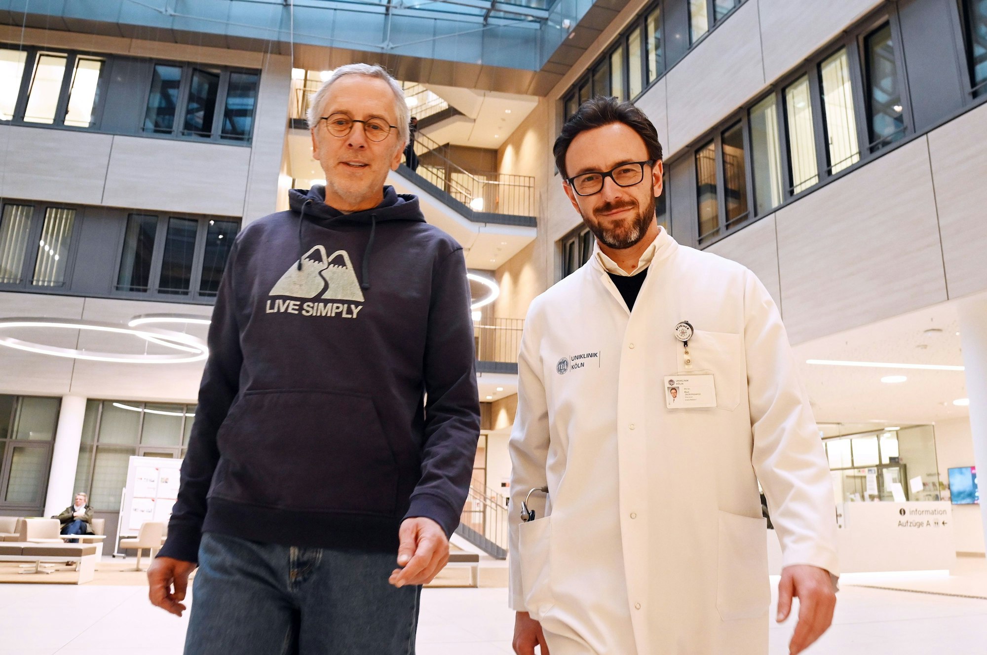 Jörg Malsam und Dr. Ron Jachimowicz sind in der Klinik zu sehen. Malsam trägt einen blauen Sweater mit dem Aufdruck "Live simply", Jachimowicz einen weißen Arztkittel.
