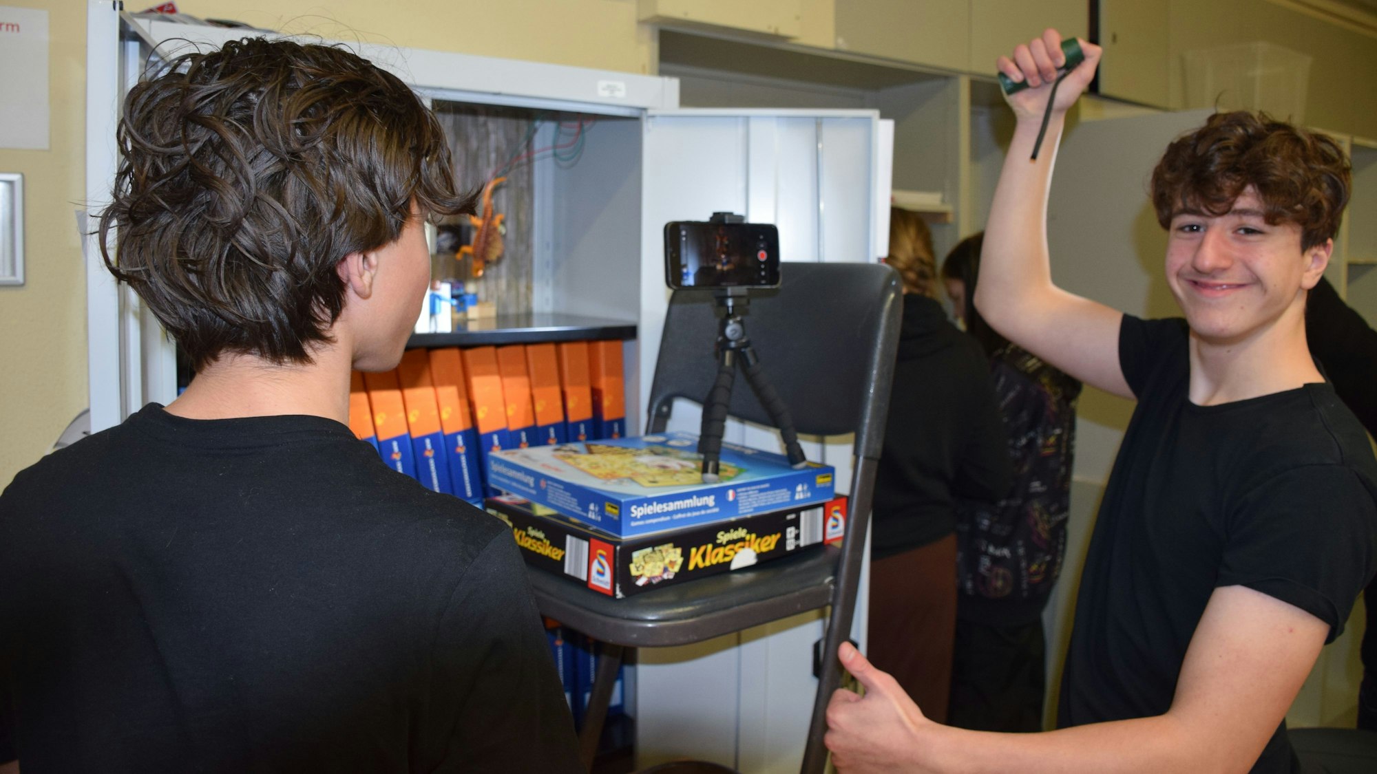 Ein Jugendlicher lächelt in die Kamera. Neben ihm ist ein Handy auf einer Halterung angebracht, die auf einem Stuhl steht, der mit zwei Brettspielverpackungen erhöht wurde.