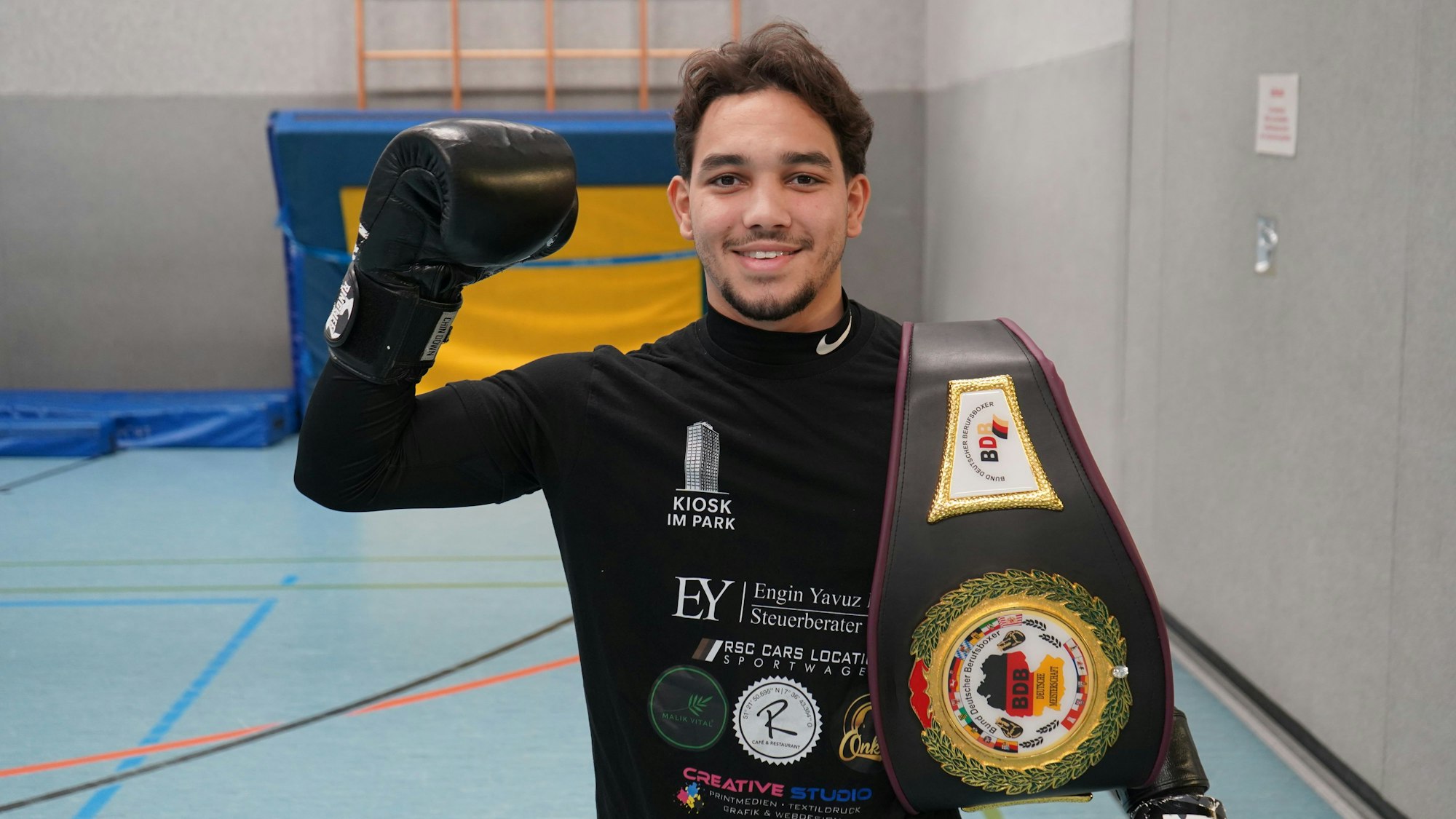 Kebdani hält eine behandschuhte Faust hoch, an seiner Schulter sein Gürtel.