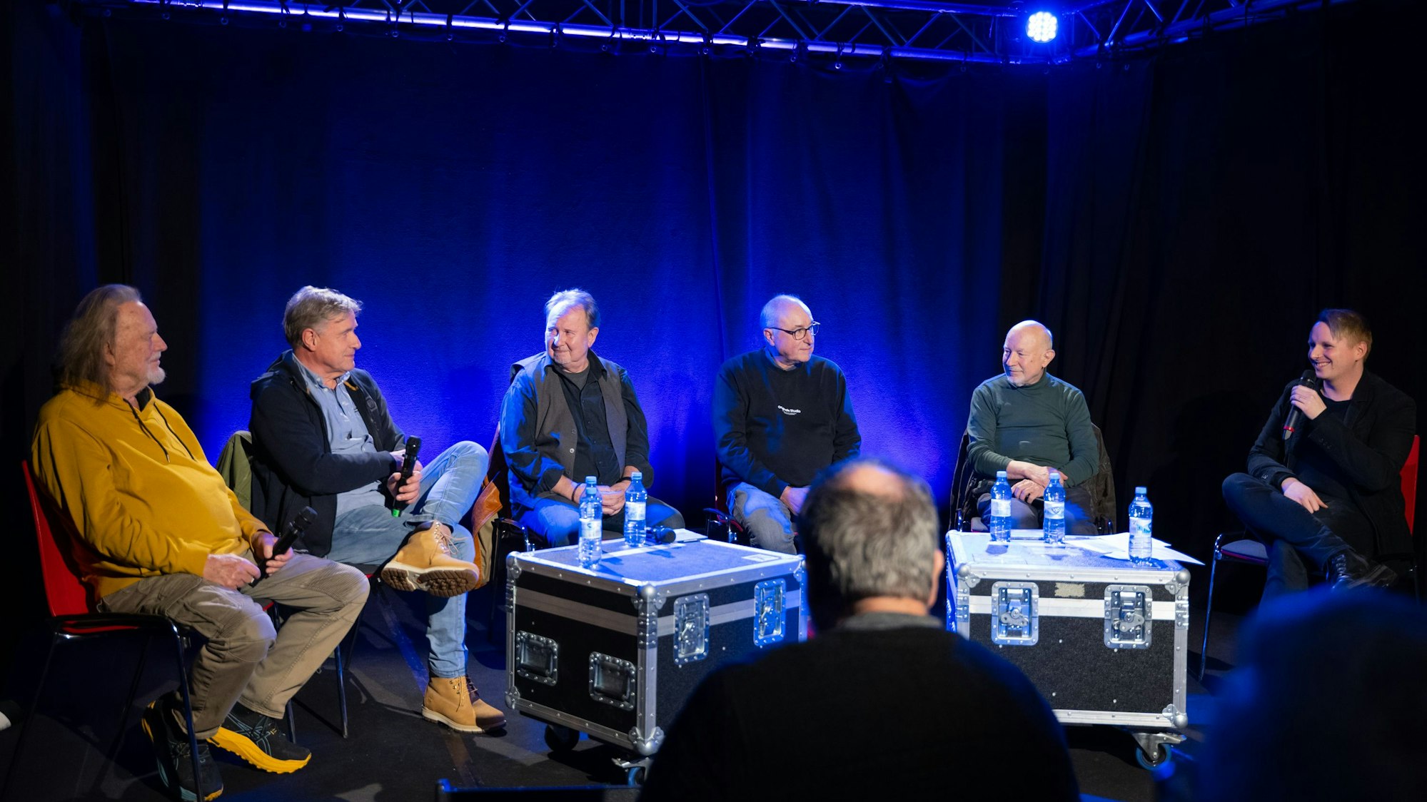 Sechs Männer sitzen nebeneinander auf einer Bühne.