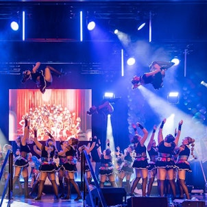 Die Aufnahme zeigt drei Tänzerinnen, die von Gruppen weiterer Tänzerinnen in die Luft geworfen werden und Salti schlagen.