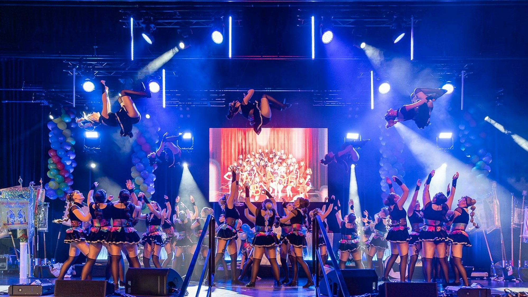 Die Aufnahme zeigt drei Tänzerinnen, die von Gruppen weiterer Tänzerinnen in die Luft geworfen werden und Salti schlagen.