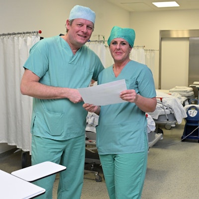 Chefarzt und OP-Managerin in OP-Kleidung stehen im Aufwachraum der Patienten und prüfen den Ausdruck für den heutigen OP-Plan.