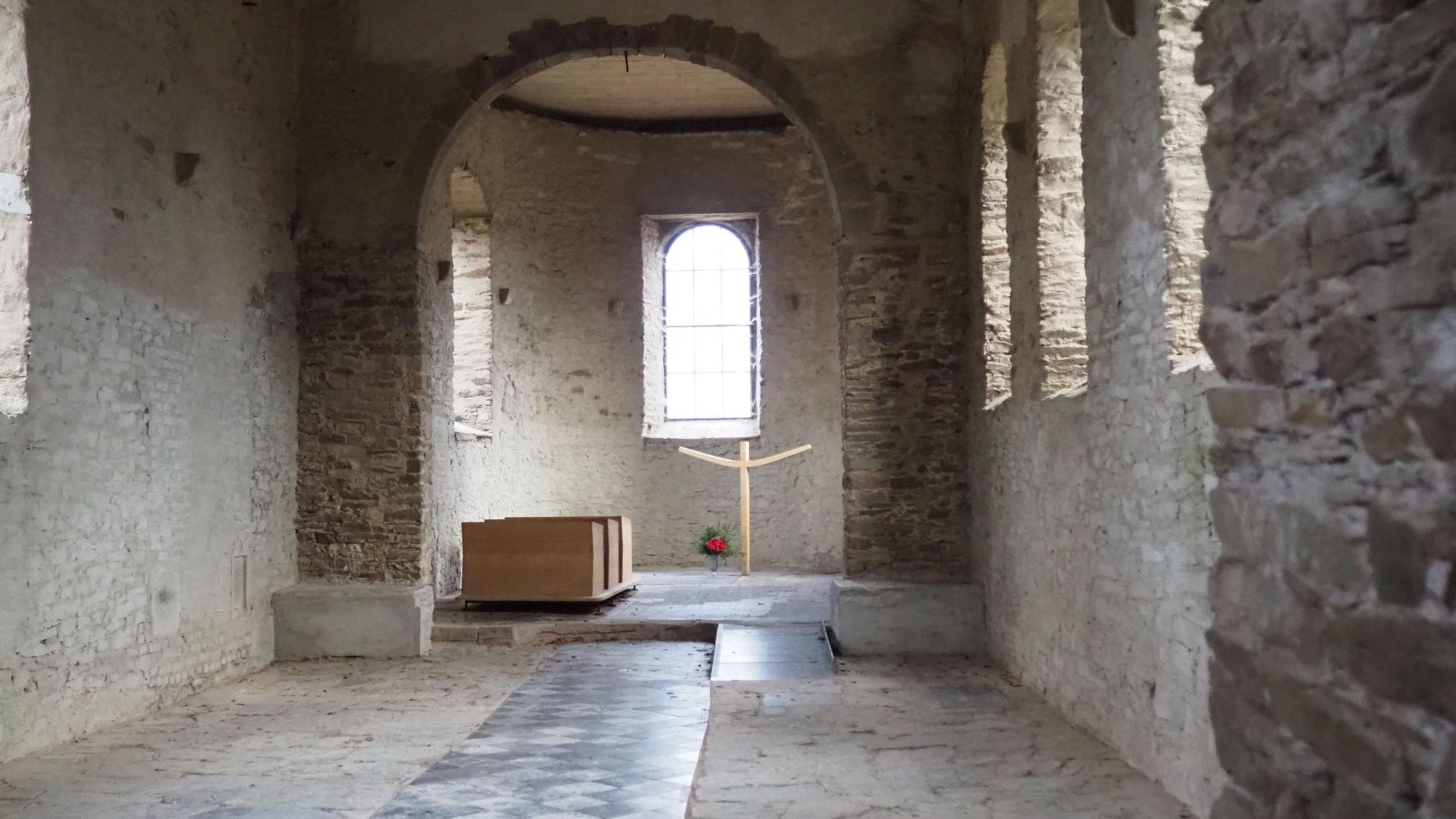 Blick in das unverputzte Kirchenschiff. Im Altarraum sind drei Sitzbänke und ein einfaches, modernes Holzkreuz zu sehen. Daneben eine Vase mit Blumen.