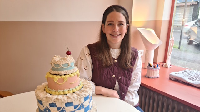 Eine junge Frau mit langem dunklem Haar sitzt an einem Tisch, auf dem ein Tortenmodell steht.