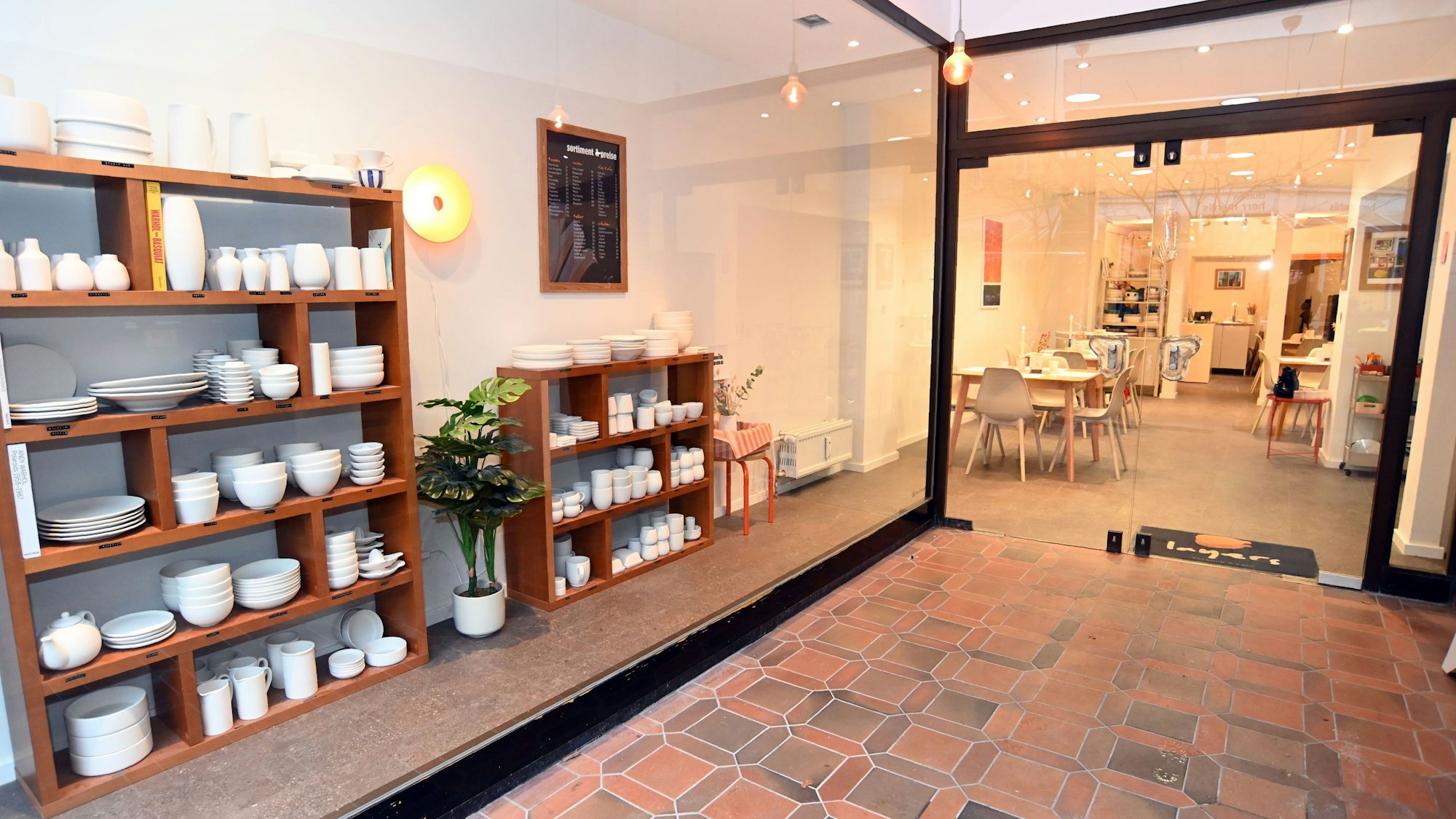 Ein Blick ins Studio von draußen. Links stehen Keramikrohlinge im Regal. Im Hauptraum stehen Tische und Stühle.