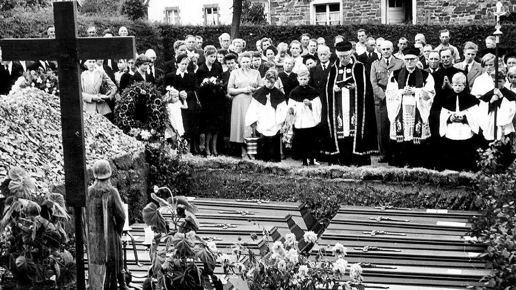 In einem Gemeinschaftsgrab sind zahlreiche Särge nebeneinander aufgebahrt worden. Vor dem Grab ein Priester und Messdiener und die Gemeinde der Wollseifener, die an der Umbettung teilnahm.