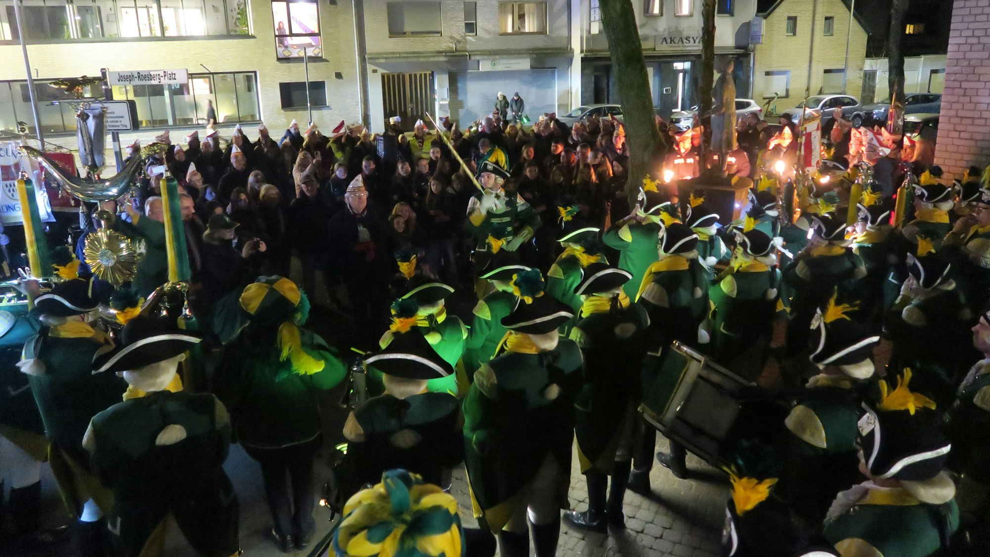 Viele Karnevalisten in Uniform und mit Musikinstrumenten stehen auf einem Platz an einem Brunnen.