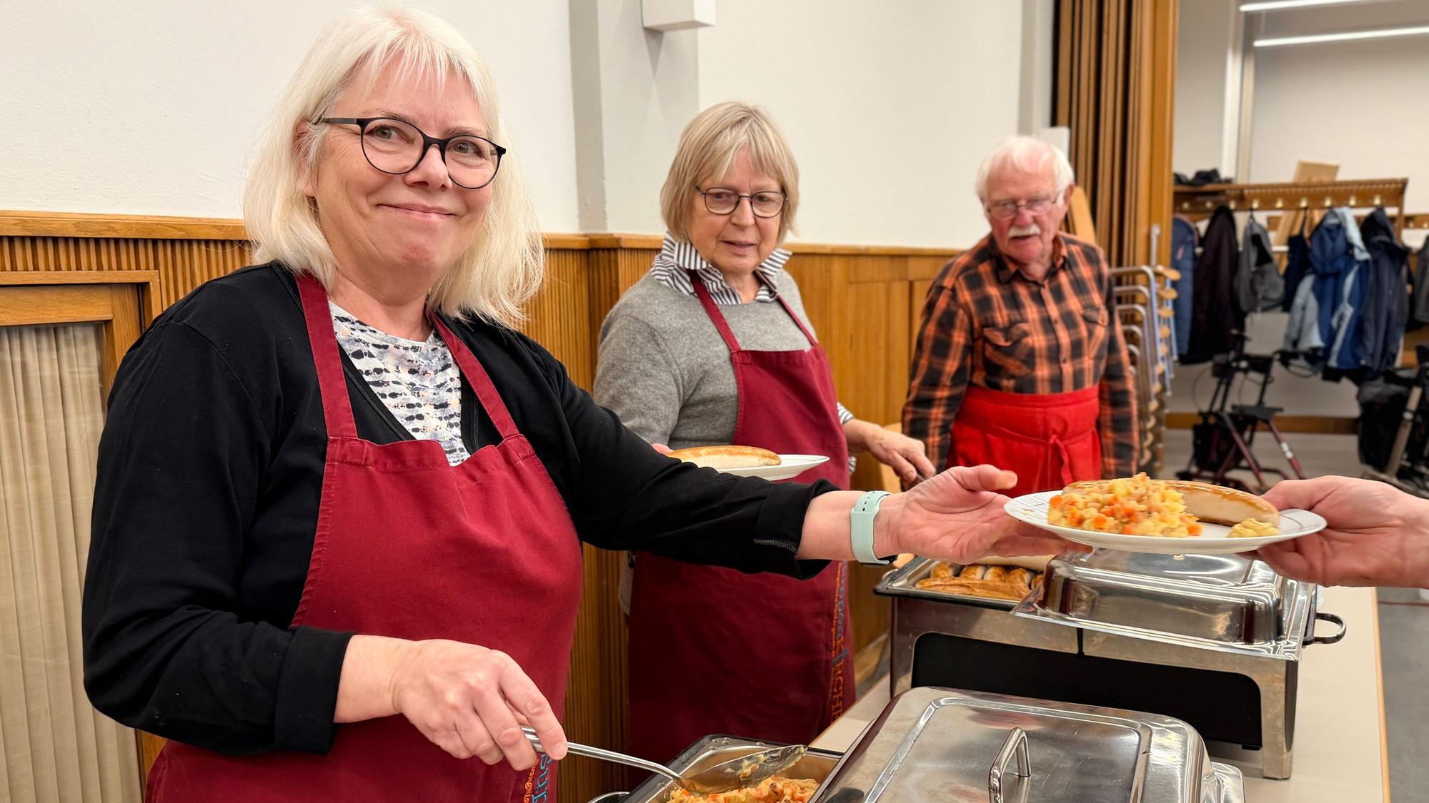 Zwei Frauen und ein Mann, die alle eine rote Schürze tragen, füllen die Teller mit dem Essen.