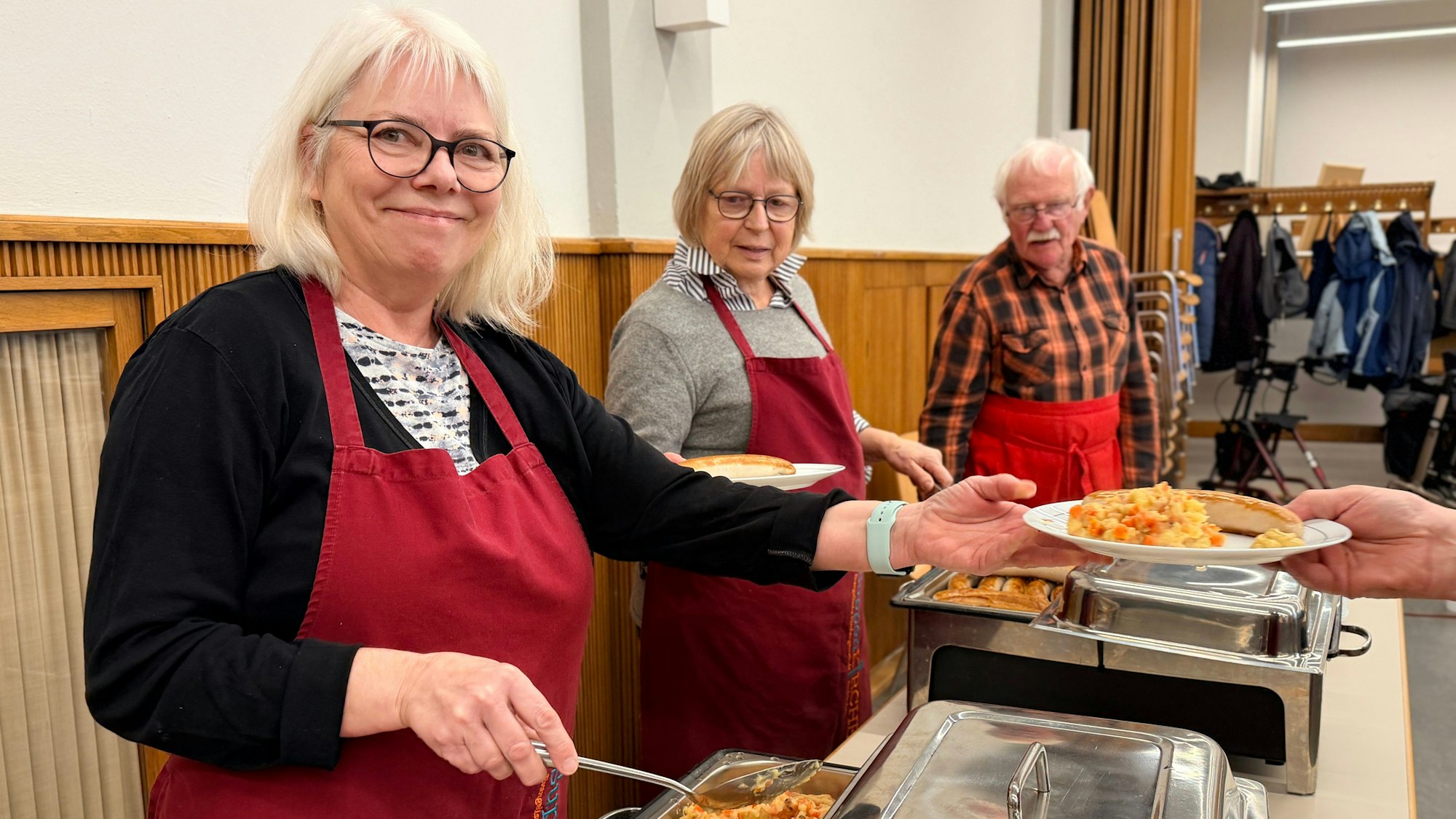 Zwei Frauen und ein Mann, die alle eine rote Schürze tragen, füllen die Teller mit dem Essen.