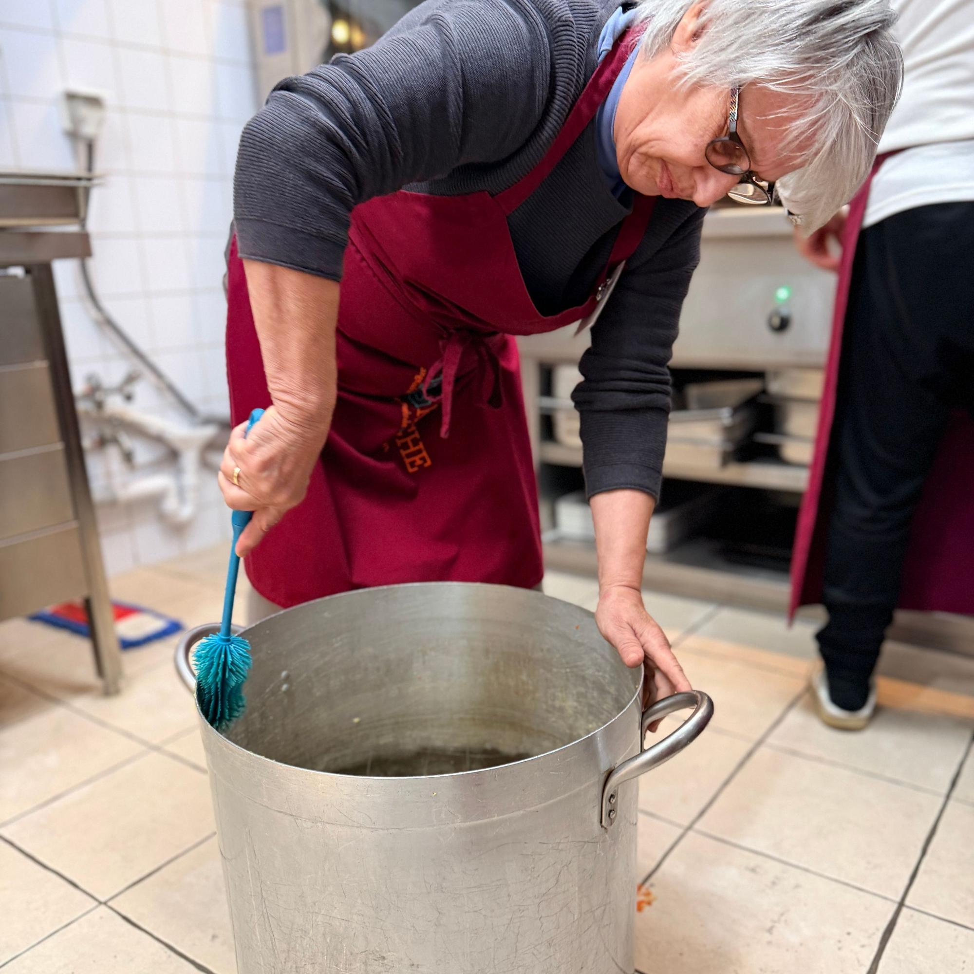 Eine Frau steht vornübergebeugt über einem großen Kochtopf, der auf dem Küchenboden steht, und schrubbt diesen mit einer Bürste sauber.