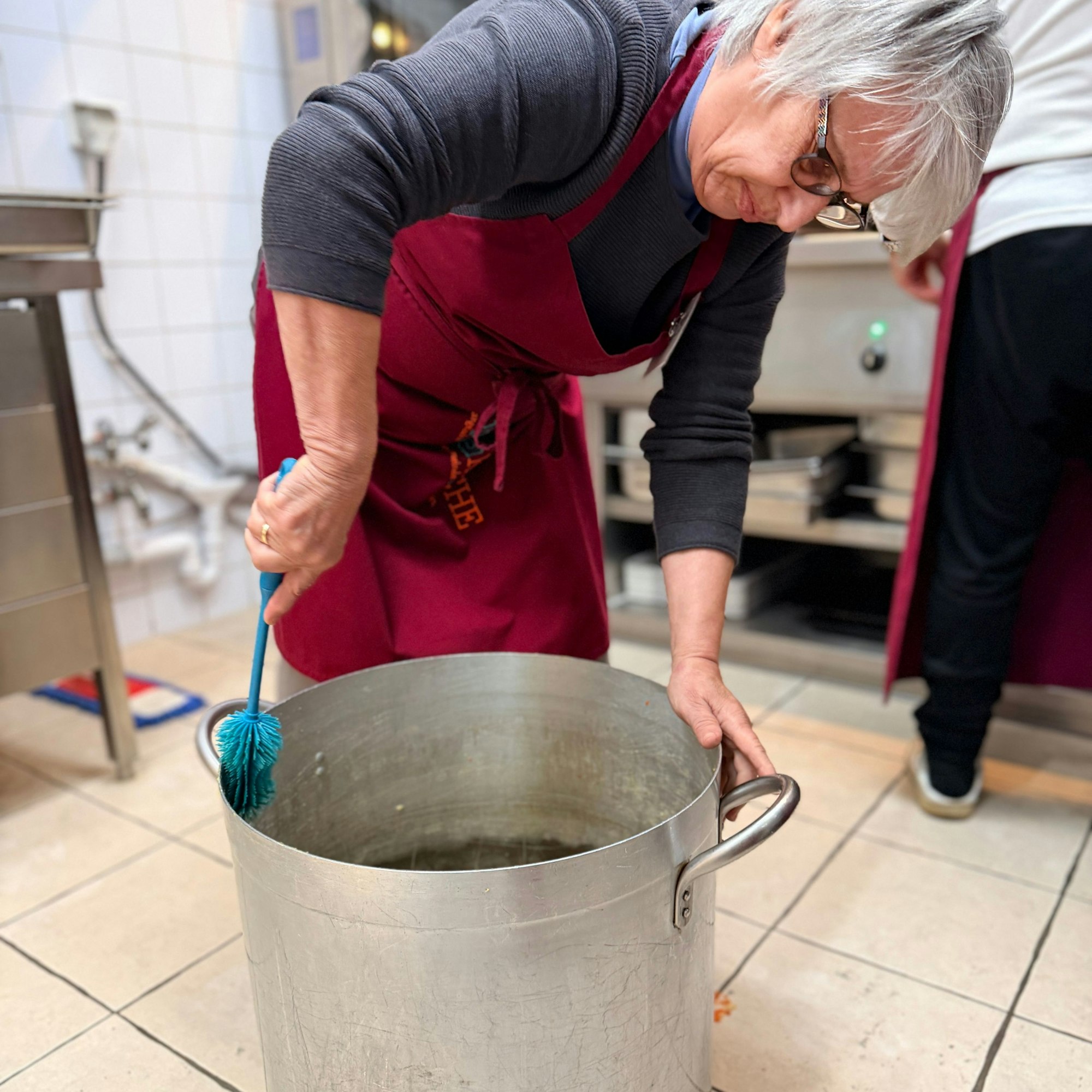 Eine Frau steht vornübergebeugt über einem großen Kochtopf, der auf dem Küchenboden steht, und schrubbt diesen mit einer Bürste sauber.