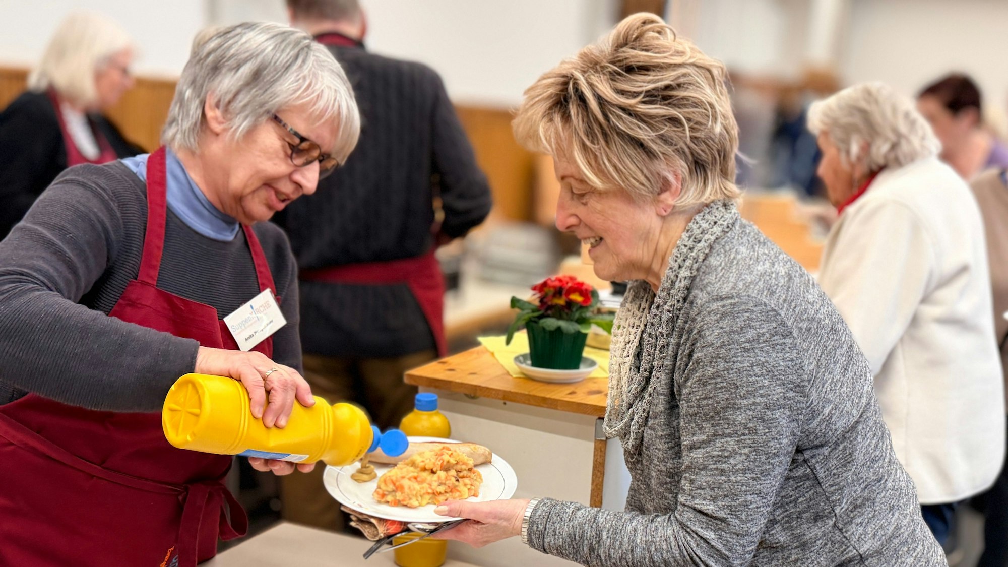 Eine der Helferinnen macht einem weiblichen Gast einen Klecks Senf auf den Teller. Dabei unterhalten sie sich.