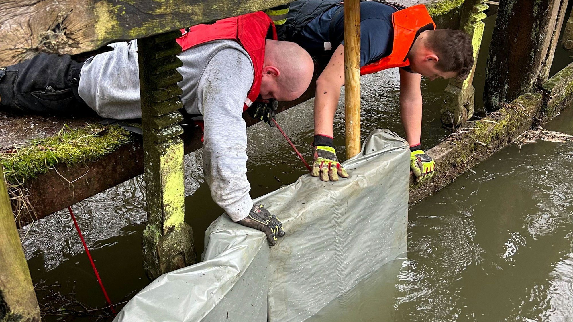 Zwei Einsatzkräfte montieren mit Folie verkleidete Paletten in den beschädigten Bereich des Absperrbauwerks vor dem Wassergraben in Rösrath-Venauen.