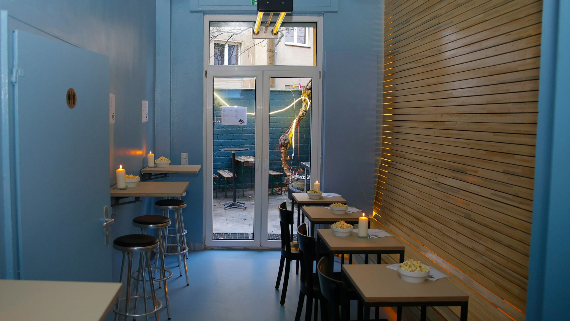Ein Blick ins Lokal auf Hinterhof durchs Fenster sowie Holztische mit Kerzen drauf im Innenraum bei der Eröffnung des Wallczka-Bistro in der Kyffhäuserstrasse 50.