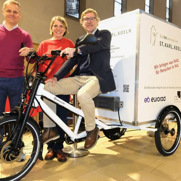 Ein Mann sitzt auf einem Lastenrad. Ein Mann und eine Frau stehen hinter ihm in einem Kirchenraum, der zur Lebensmittelausgabe umfunktioniert wurde.