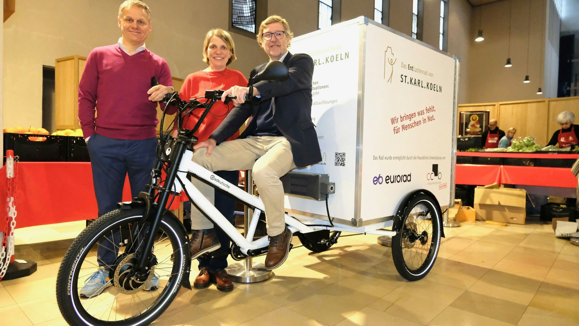 Ein Mann sitzt auf einem Lastenrad. Ein Mann und eine Frau stehen hinter ihm in einem Kirchenraum, der zur Lebensmittelausgabe umfunktioniert wurde.