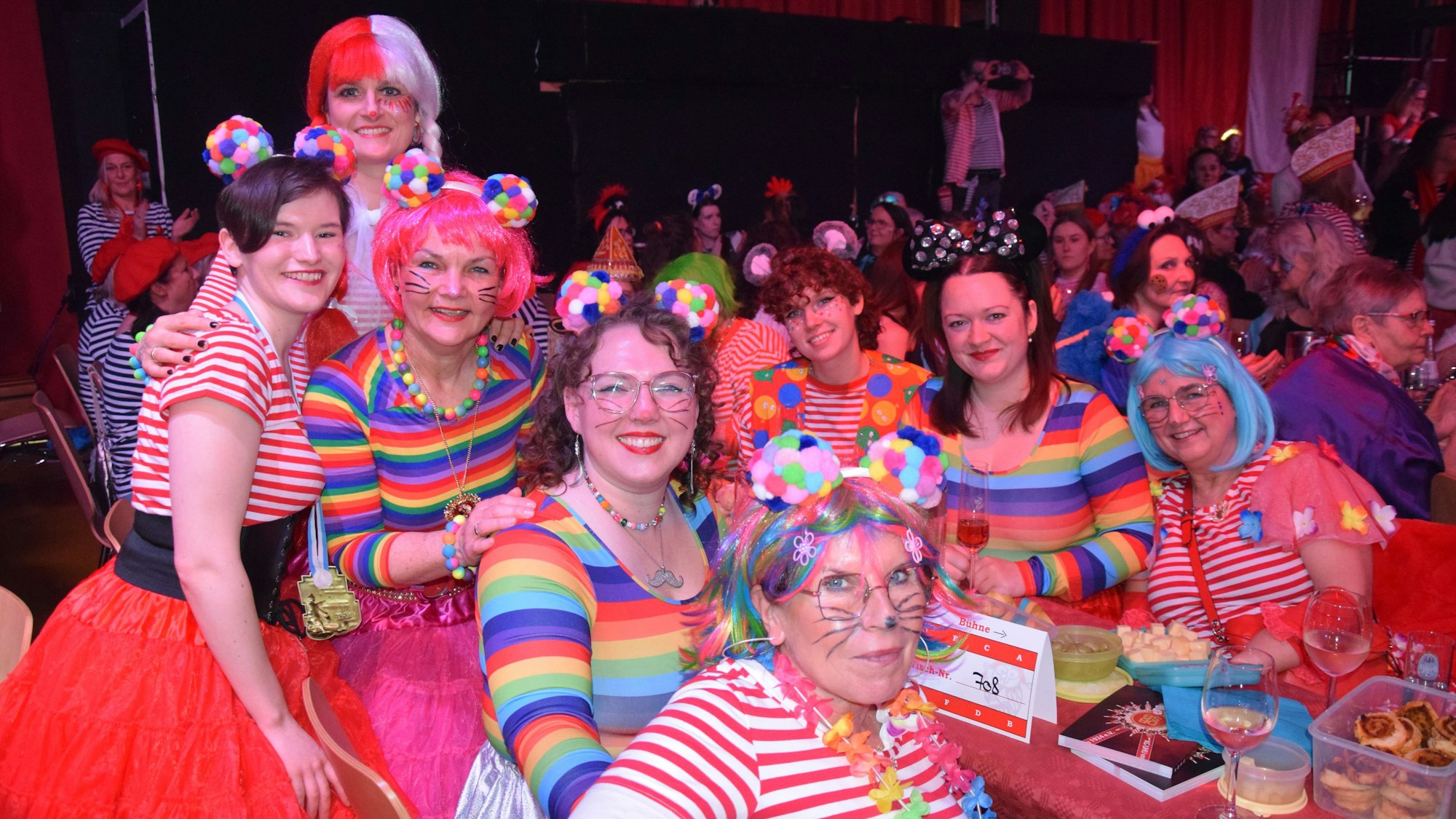 Ein Gruppe Frauen in gestreiften Oberteilen und angemalten Gesichtern sitzt an einem Biertisch in einem vollbesetzten Saal.