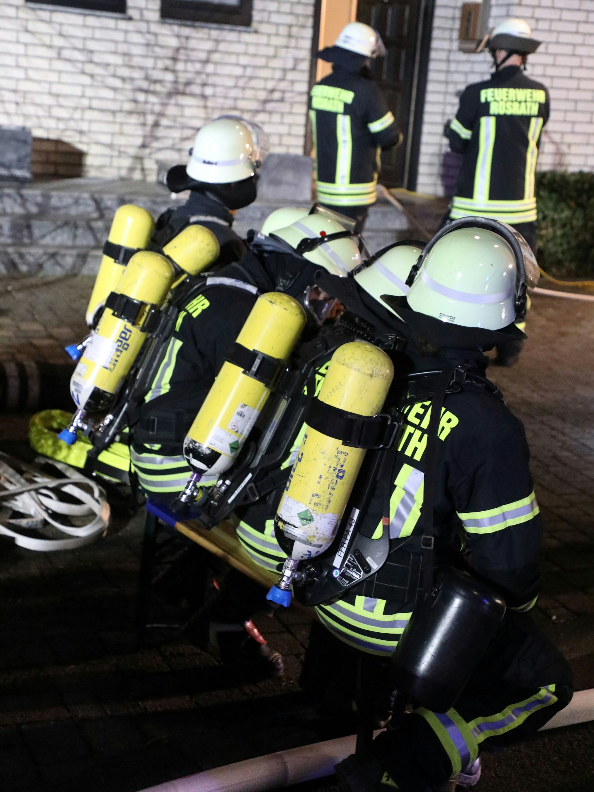Feuerwehrleute mit Pressluftatmern auf dem Rücken sitzen auf einer Bank vor einem Brandhaus.