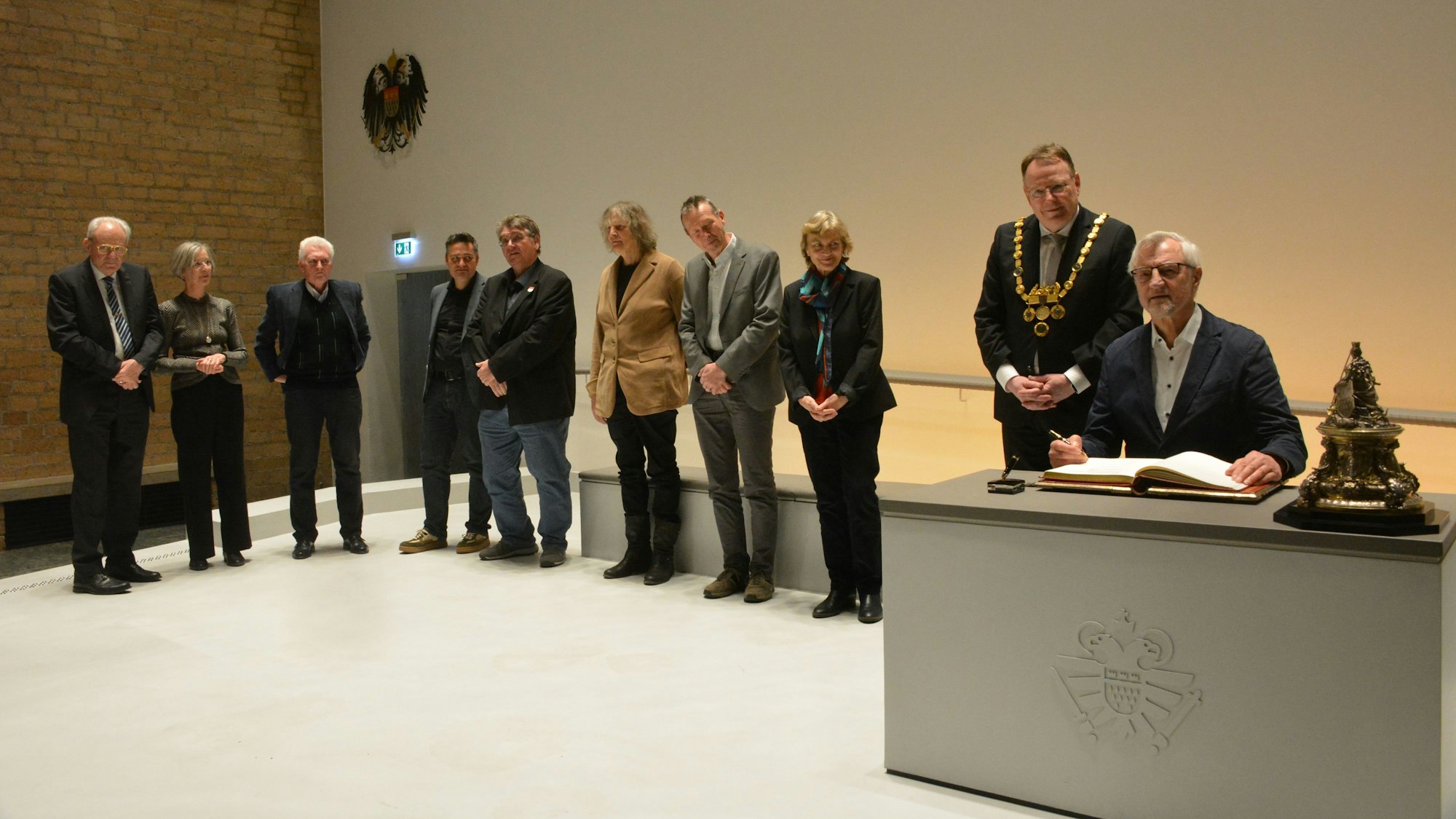 Ein Gruppe von acht Menschen steht in einer Reihe, ein Mann sitzt an einem Tisch, vor ihm liegt das Goldene Buch.