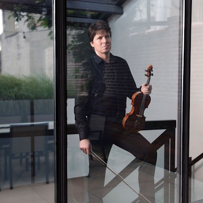 Joshua Bell posiert mit Violine und Bogen hinter einem bodentiefen Fenster.