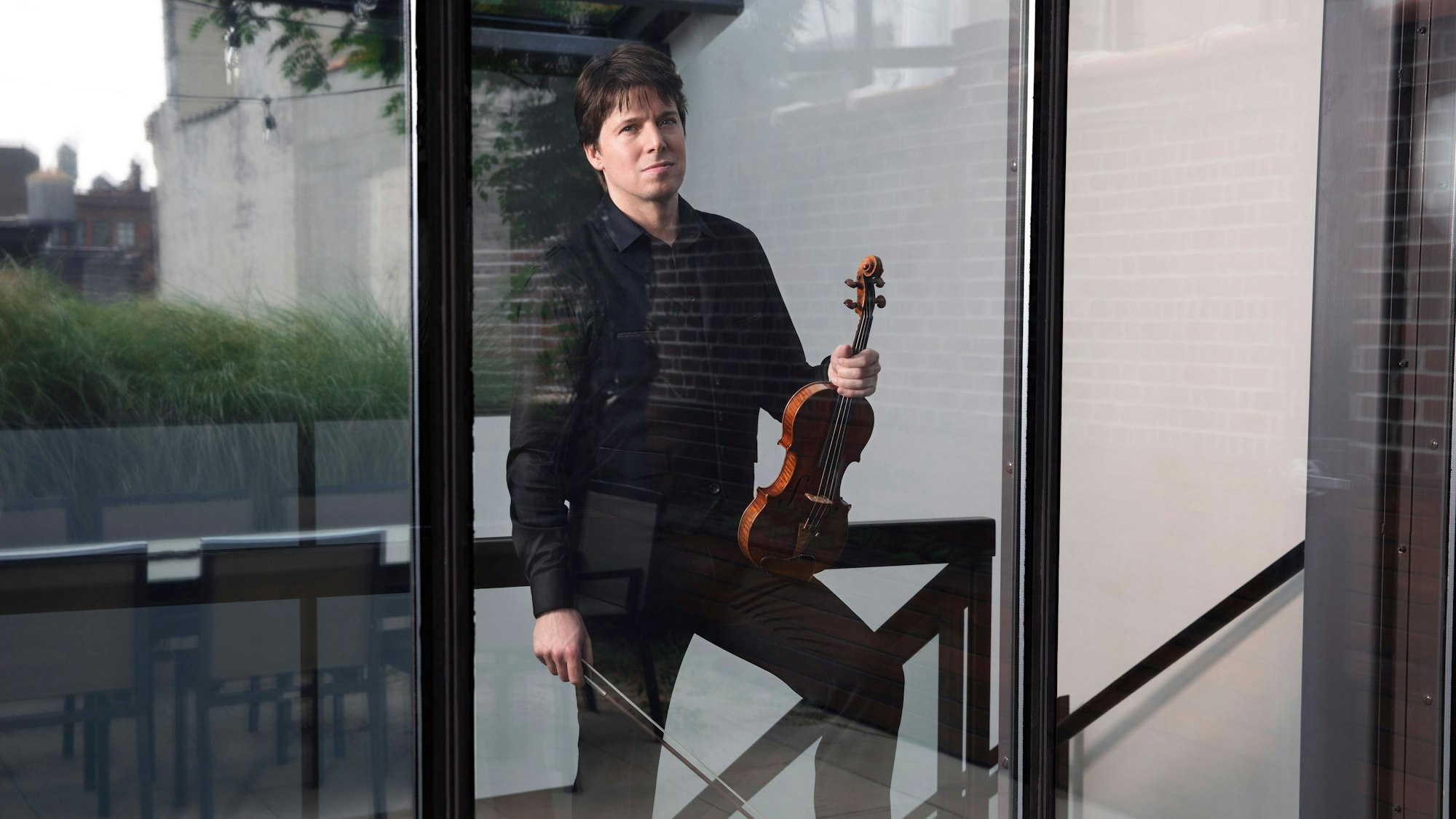 Joshua Bell posiert mit Violine und Bogen hinter einem bodentiefen Fenster.