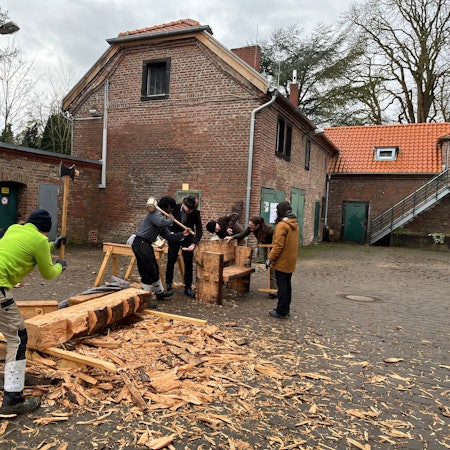 Im Innenhof eines Gutshofs arbeiten junge Leute an einem Baumstamm.