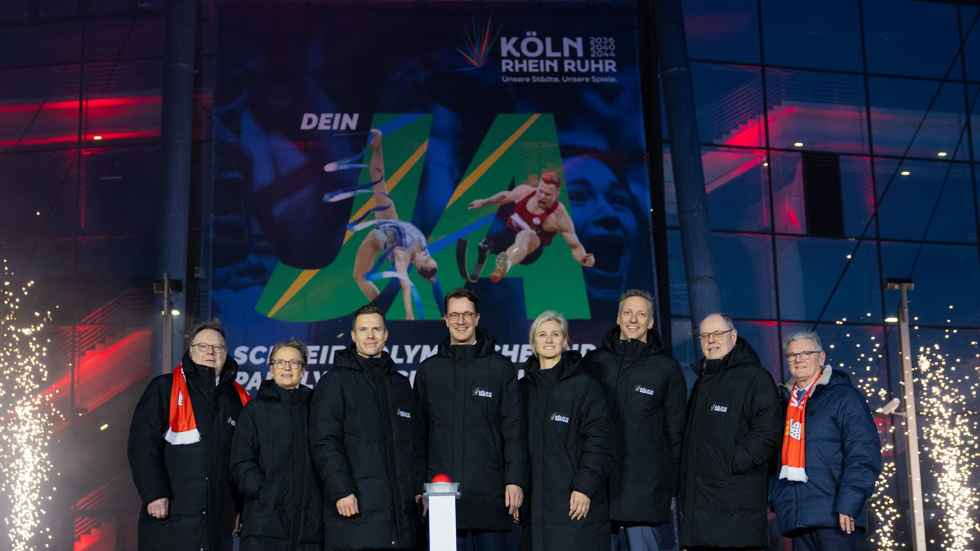 Acht Menschen stehen nebeneinander hinter einem Buzzer.