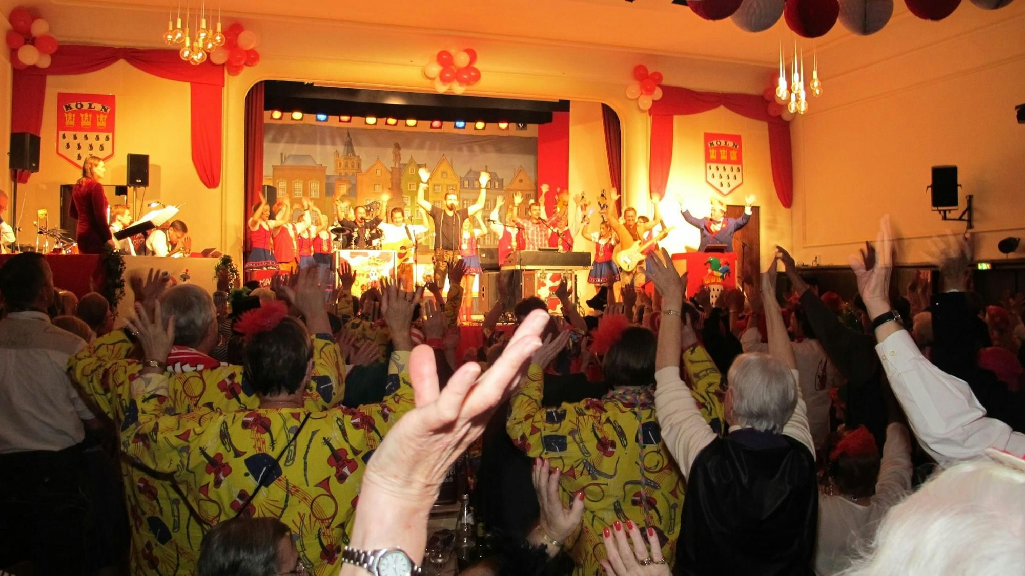 Auf der Bühne in einem Saal steht eine Band, das Publikum ist aufgestanden und streckt die Arme nach oben.