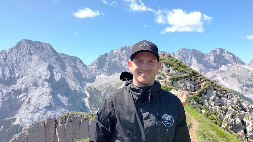 Ein junger Mann mit Kappe auf dem Kopf steht vor einem Bergpanorama.