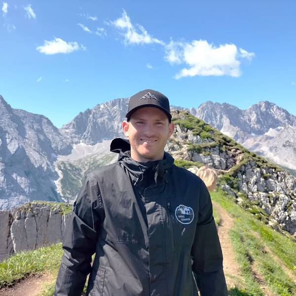 Ein junger Mann mit Kappe auf dem Kopf steht vor einem Bergpanorama.