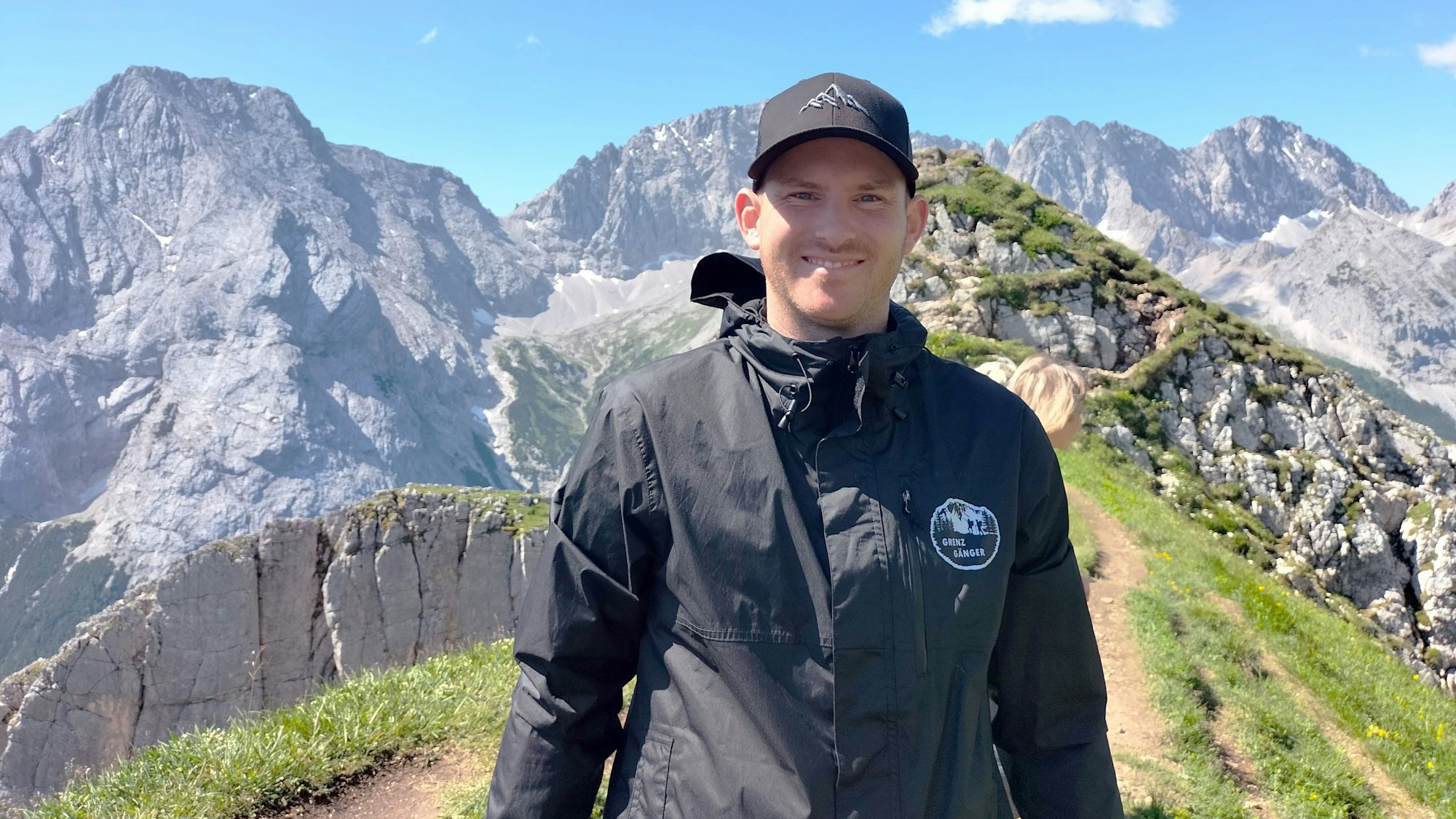 Ein junger Mann mit Kappe auf dem Kopf steht vor einem Bergpanorama.