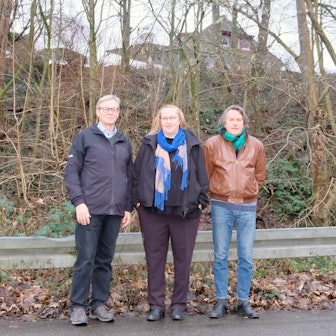 Zwei Männer und eine Frau stehen am Straßenrand, hinter ihnen ragt einen mit Bäumen bewachsener Hang auf.