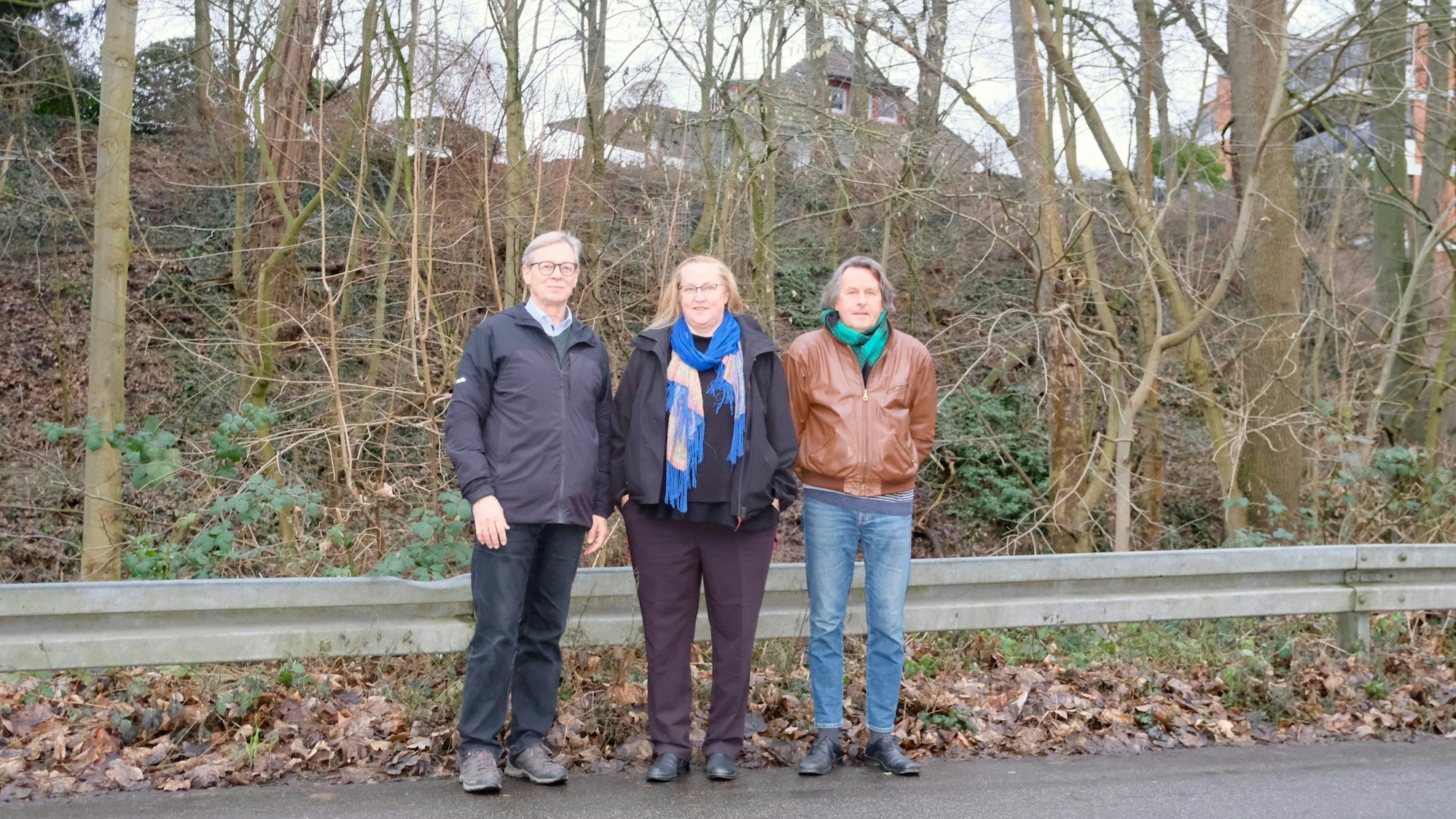 Zwei Männer und eine Frau stehen am Straßenrand, hinter ihnen ragt einen mit Bäumen bewachsener Hang auf.