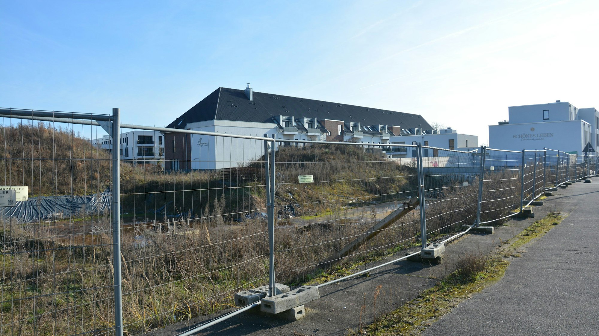 Zu sehen ist ein Bauzaun vor einer Baugrube, dahinter ein Gebäudekomplex.