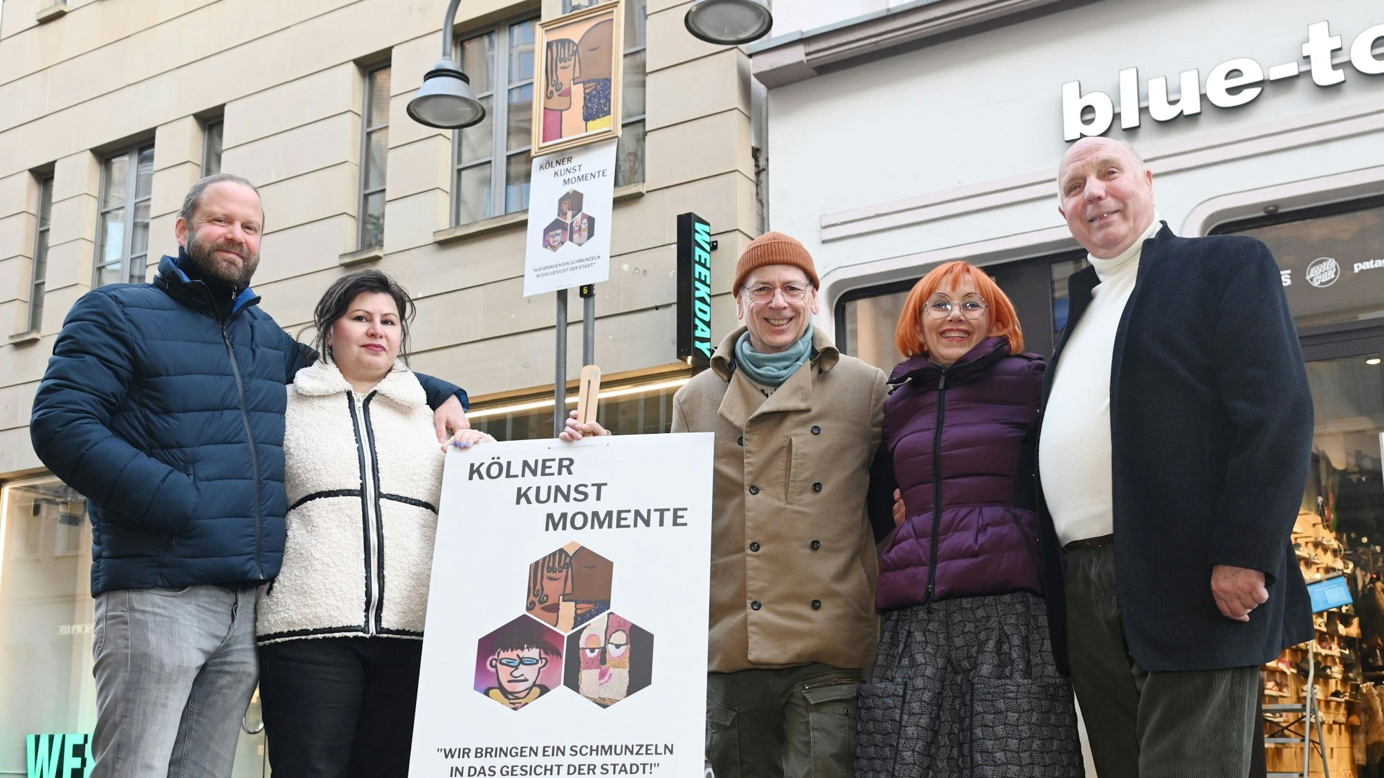 Aktion „Kölner Kunst Momente“ auf der Ehrenstraße: Mit Moderator Thomas Hackenberg (m.) ("Quiz Taxi") alias Thomas von Klettenberg, Kurator Roderik Helms (l.), Jean Nejadi (2.v.l. ) und Künstlerin Lucie Albrecht (2.v.r.) und Peter Möltgen von der Parfümerie Möltgen. Bunte Gesichter, auf A1-Plakate gedruckt, werden an Laternenkronen auf der Ehrenstraße angebracht.