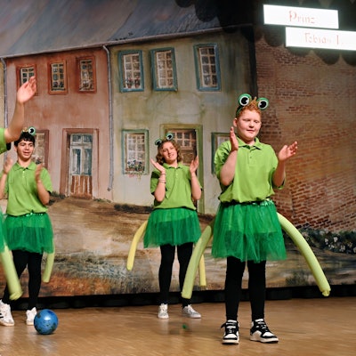 Kinder in Froschkostümen klatschen auf einer Bühne in die Hände.