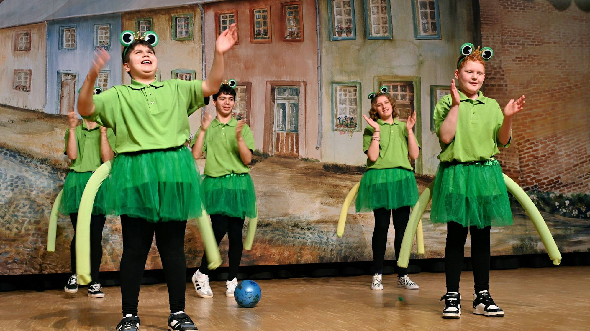 Kinder in Froschkostümen klatschen auf einer Bühne in die Hände.
