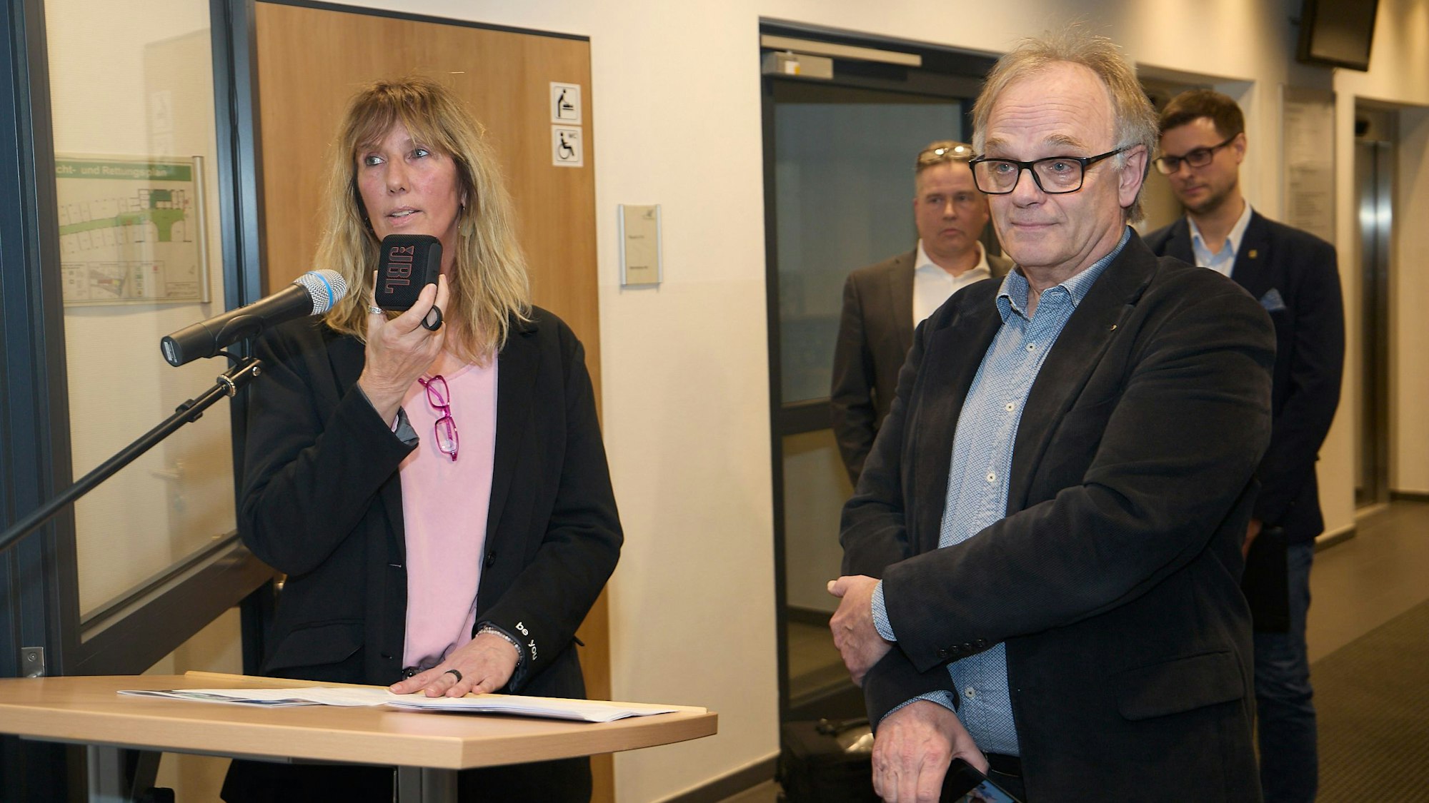 Kuratorin Ela Rübenach hält ein Aufnahmegerät an ein Mikrofon. Nebn ihr steht farrer Dr. Michael Stöhr, der mit seiner linken Hand an seinen rechten Arm fasst.