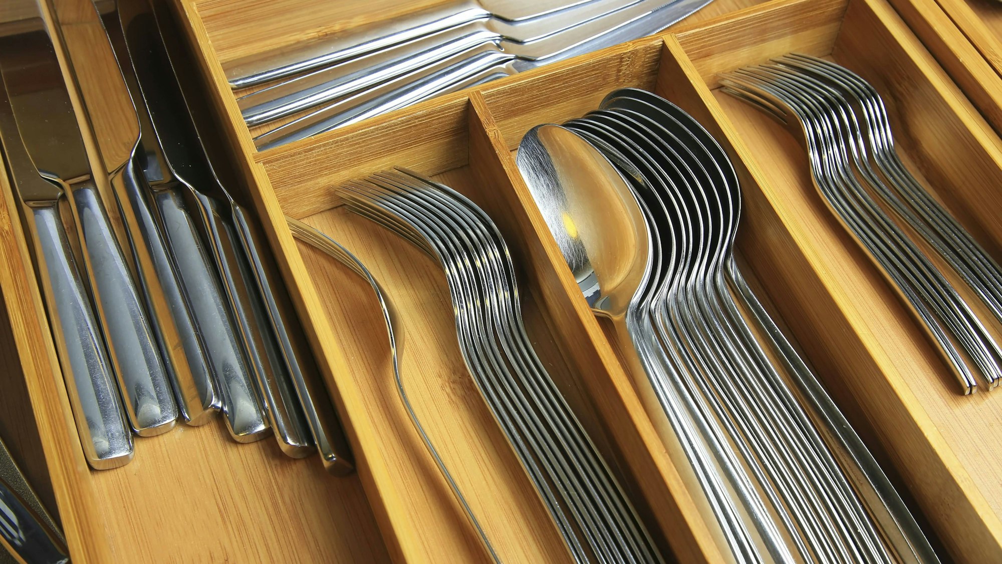Holzeinsatz in einer Besteckschublade mit mehreren Fächern, in denen Messer, Gabeln und Löffel aus Edelstahl sauber und parallel geordnet sind.