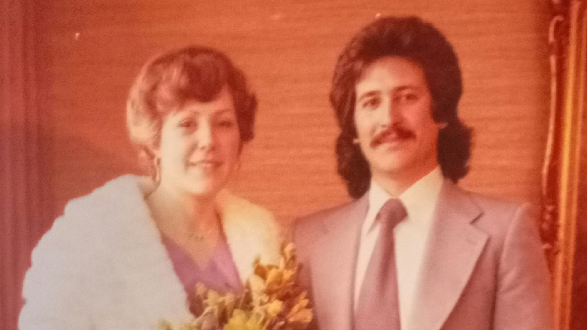 Elisabeth Capua trägt ein violettes Kleid und einen weißen Mantel, sie hält Blumen. Mario Capua trägt einen Anzug mit Krawatte.