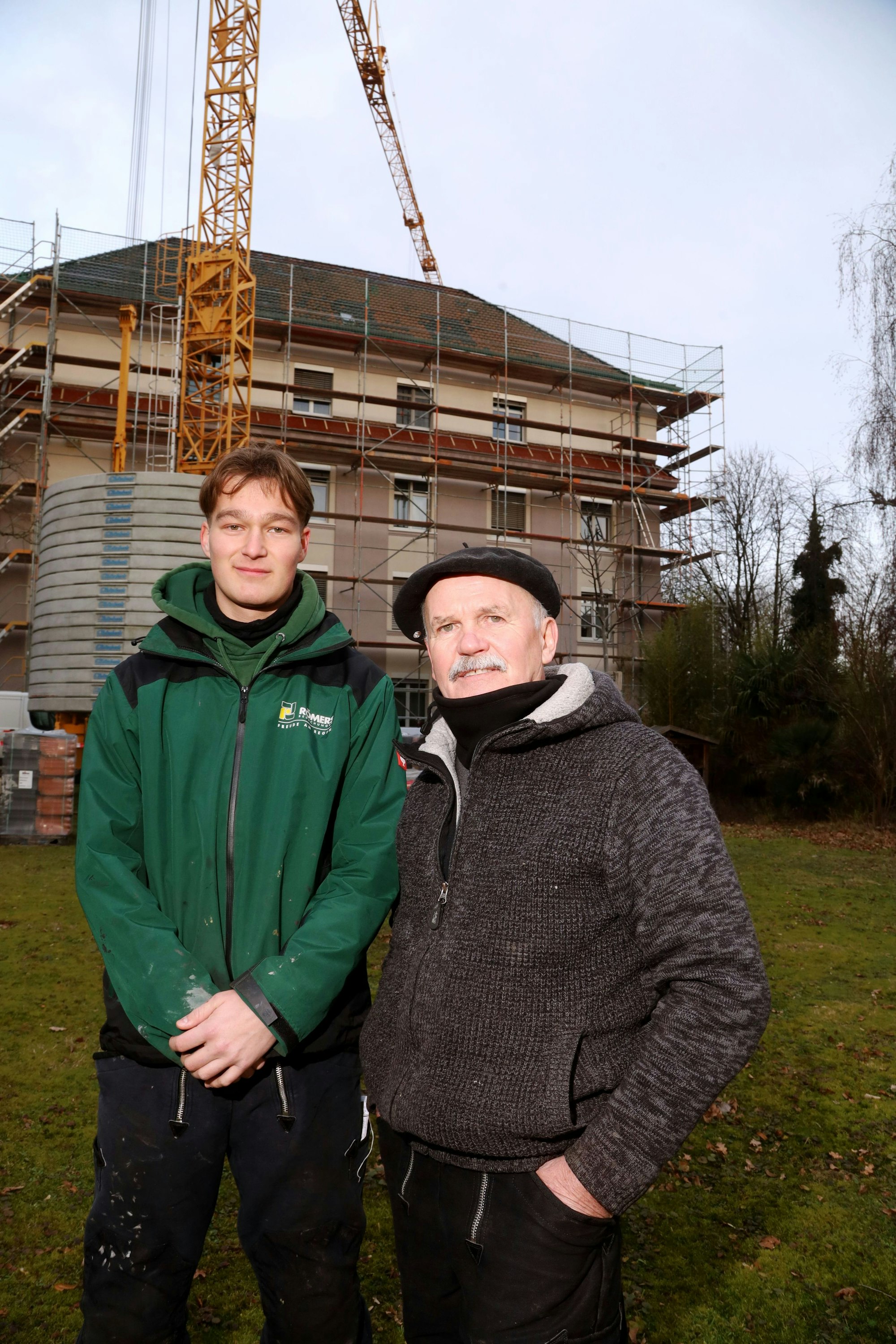 Senior-Dachdecker Bernhard Bartnik ist mit Geselle Noah Beckers zu sehen vor einem eingerüsteten Haus.