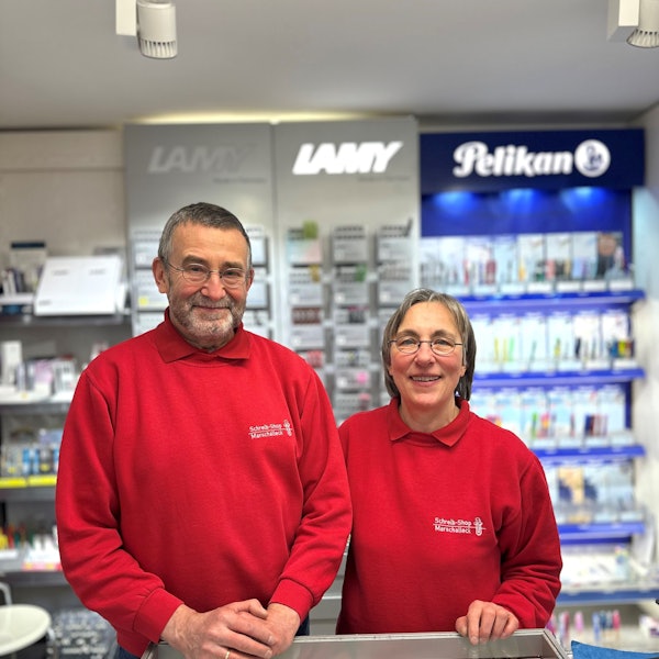 Eine Frau und ein Mann in roten Pullovern stehen vor einer Wand mit Schreibwaren.