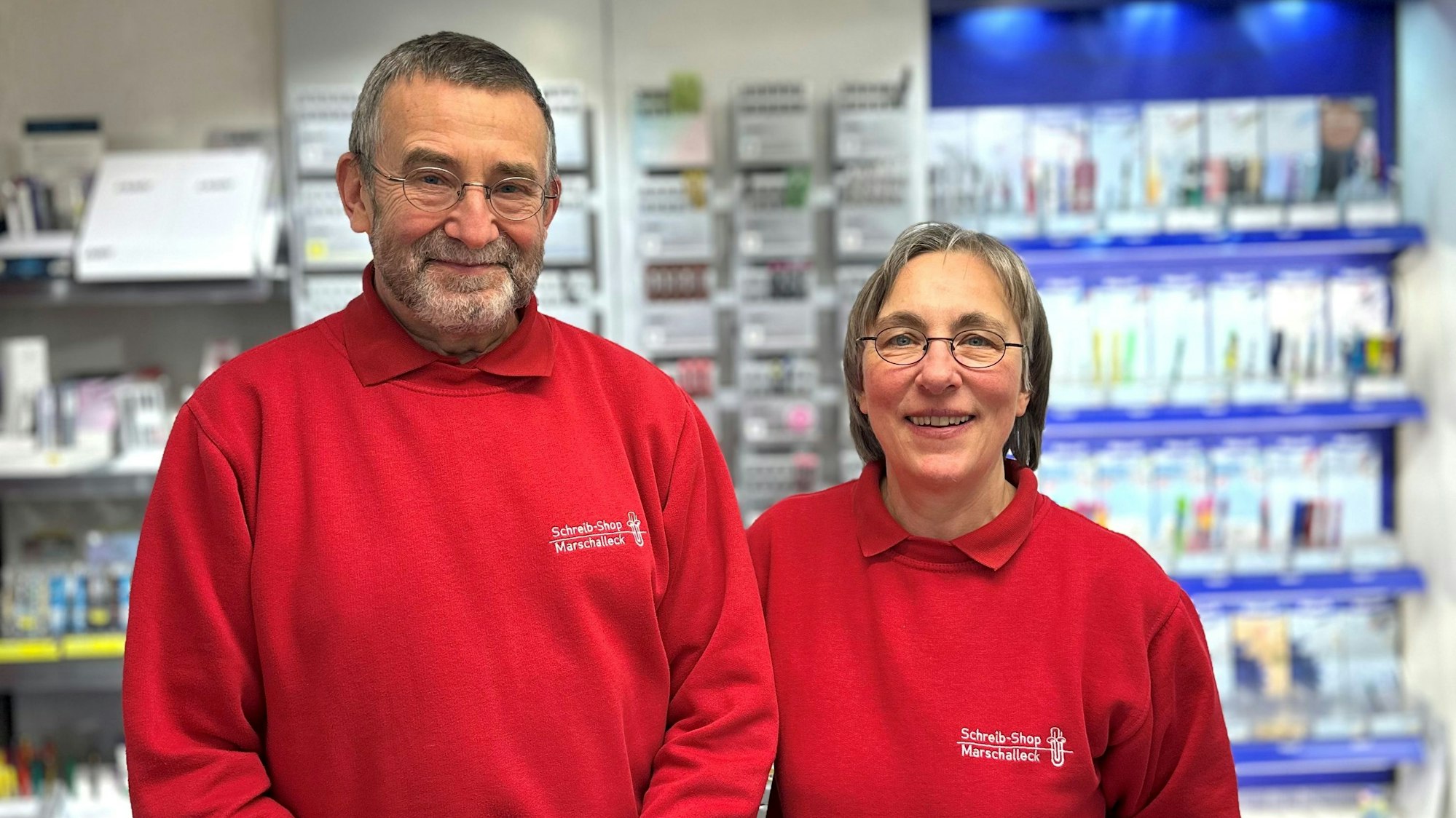 Eine Frau und ein Mann in roten Pullovern stehen vor einer Wand mit Schreibwaren.