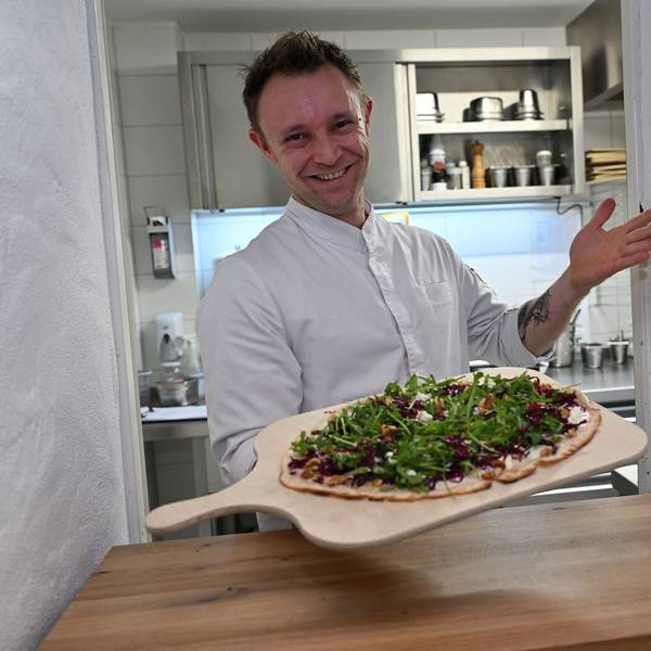 Ein lachender Koch in weißer Jacke reicht dem Betrachter einen Flammkuchen mit grünem Salat auf einem großen Holzbrett entgegen.