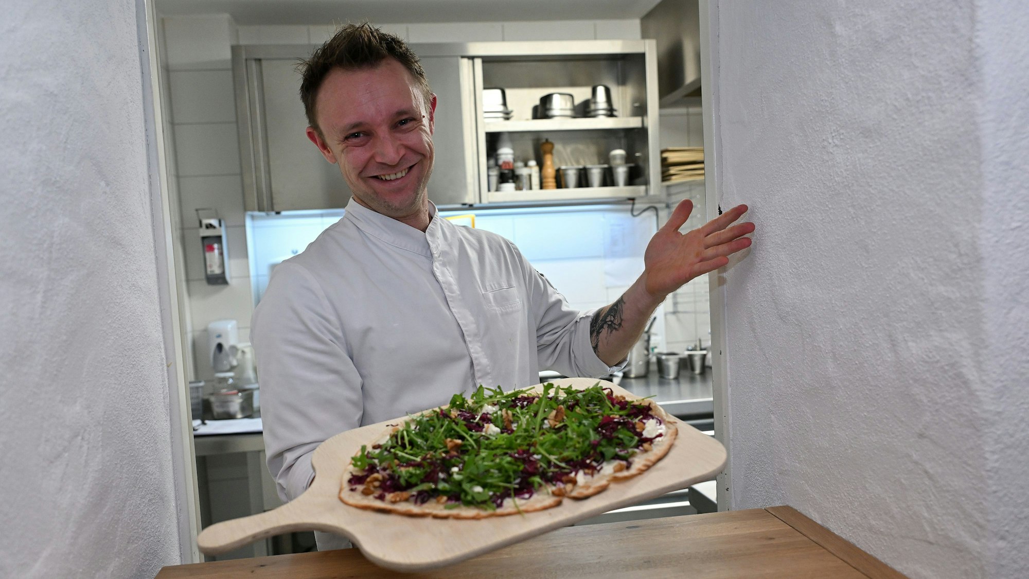 Ein lachender Koch in weißer Jacke reicht dem Betrachter einen Flammkuchen mit grünem Salat auf einem großen Holzbrett entgegen.