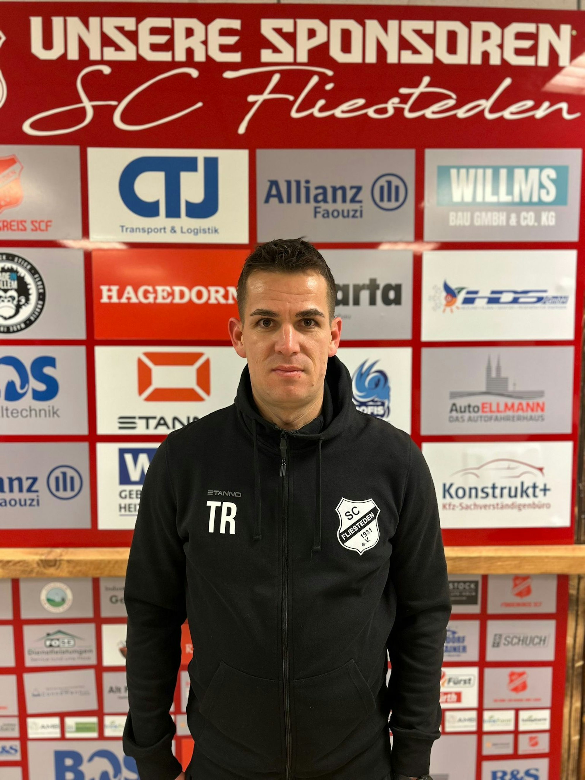 Das Bild zeigt Helge Hohl, neuer Trainer des SCF, vor der Sponsorenwand des SC Fliesteden.