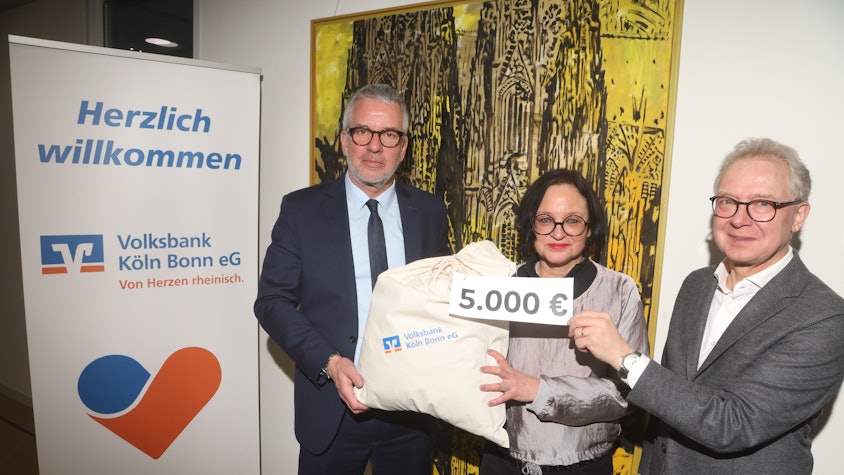 Jürgen Pütz, Vorstandsvorsitzender der Volksbank Köln Bonn, überreicht vor einem modernen Gemälde des Kölner Doms Christian Hümmeler und Caroline Kron vom „wir helfen“-Vorstandsteam einen symbolischen Spendensack über 5.000 Euro.